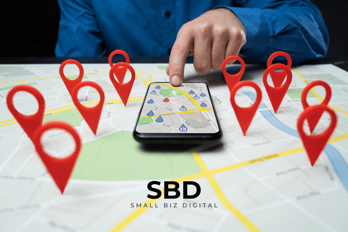 A close-up of a person's hand using a smartphone map app, surrounded by large physical red map pins standing upright on a flat paper map.
